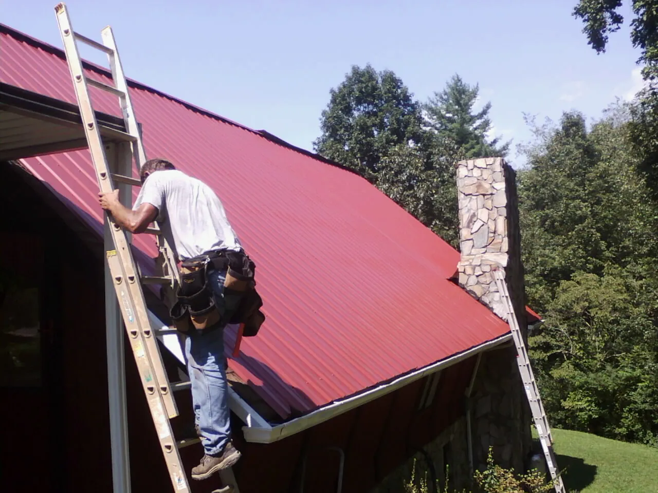 Roofer on a ladder beside a red metal roof and stone chimney — Soffit & Fascia Repair by Roofing Brothers in Cottage Grove